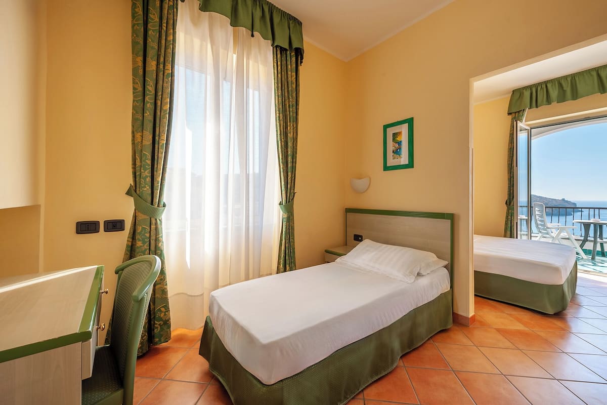 A hotel room with a bed, a chair, a small table, and a view of the sea.