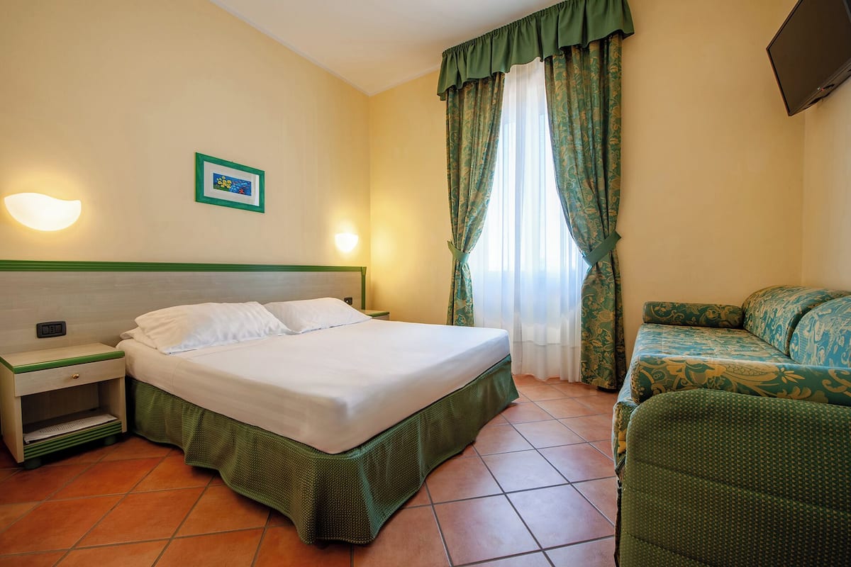 A hotel room with a bed, a nightstand, a green sofa, a TV, and a window with curtains.