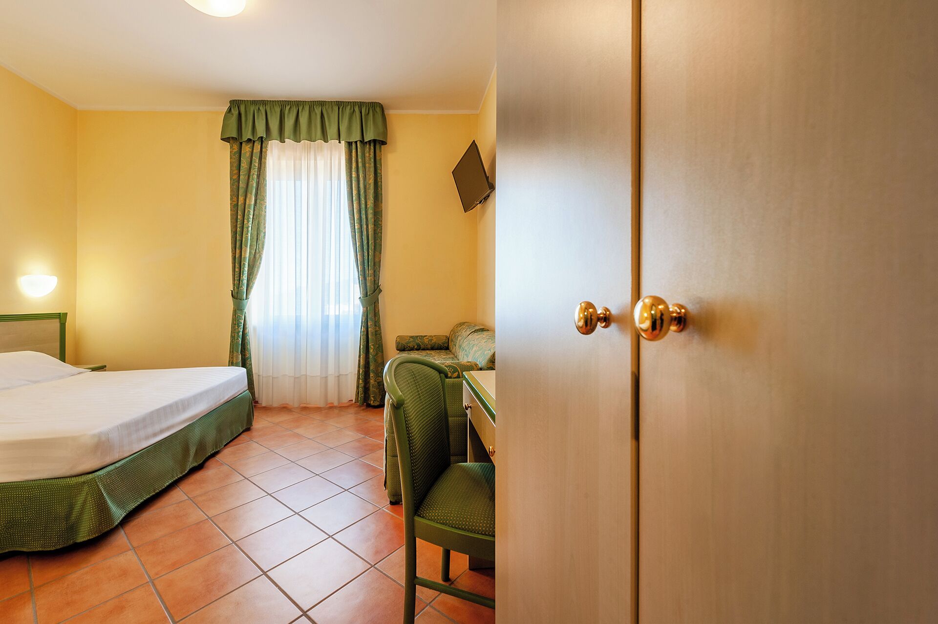 A hotel room with a bed, a chair, a desk, a TV, and a door.