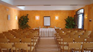 Un salón de conferencias con hileras de sillas de madera, una mesa blanca con una pantalla para proyector y dos plantas en macetas.