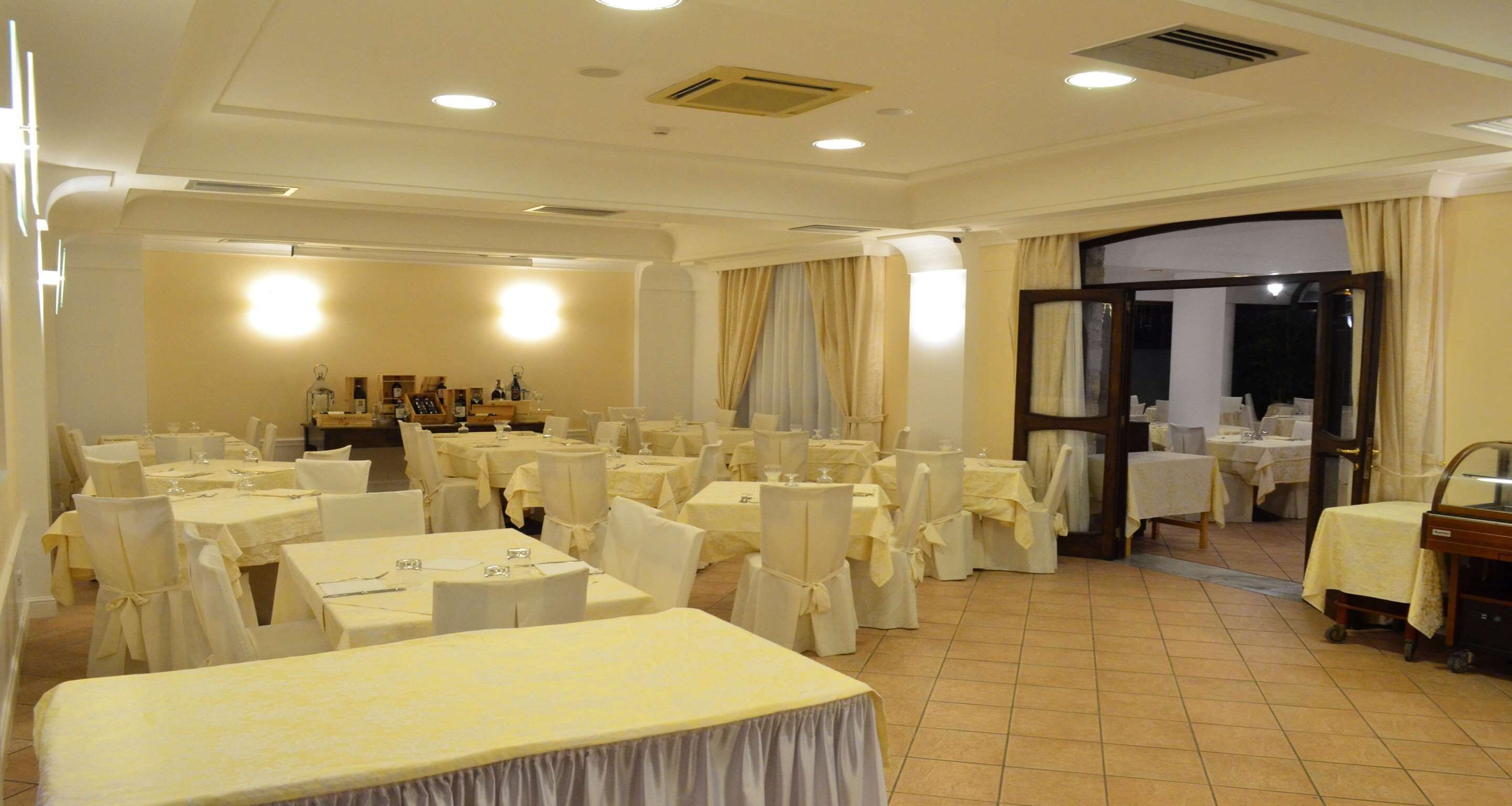 Un salón de banquetes con varias mesas redondas y sillas, una zona de recepción con mostrador, y una puerta que da al exterior.