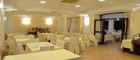 Un salón de banquetes con varias mesas redondas y sillas, una zona de recepción con mostrador, y una puerta que da al exterior.