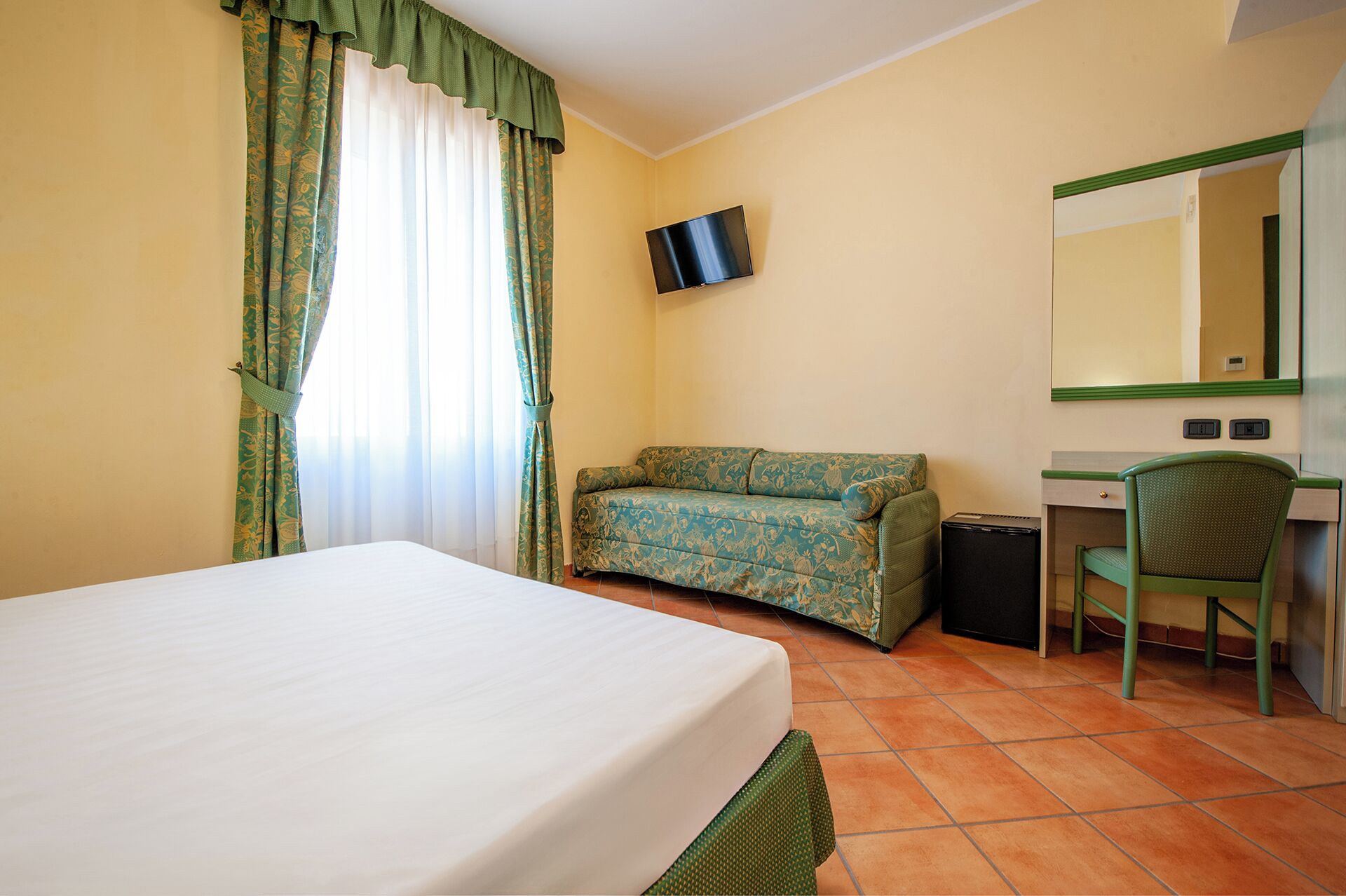 A hotel room with a bed, a sofa, a desk, a chair, a TV, and a mirror.