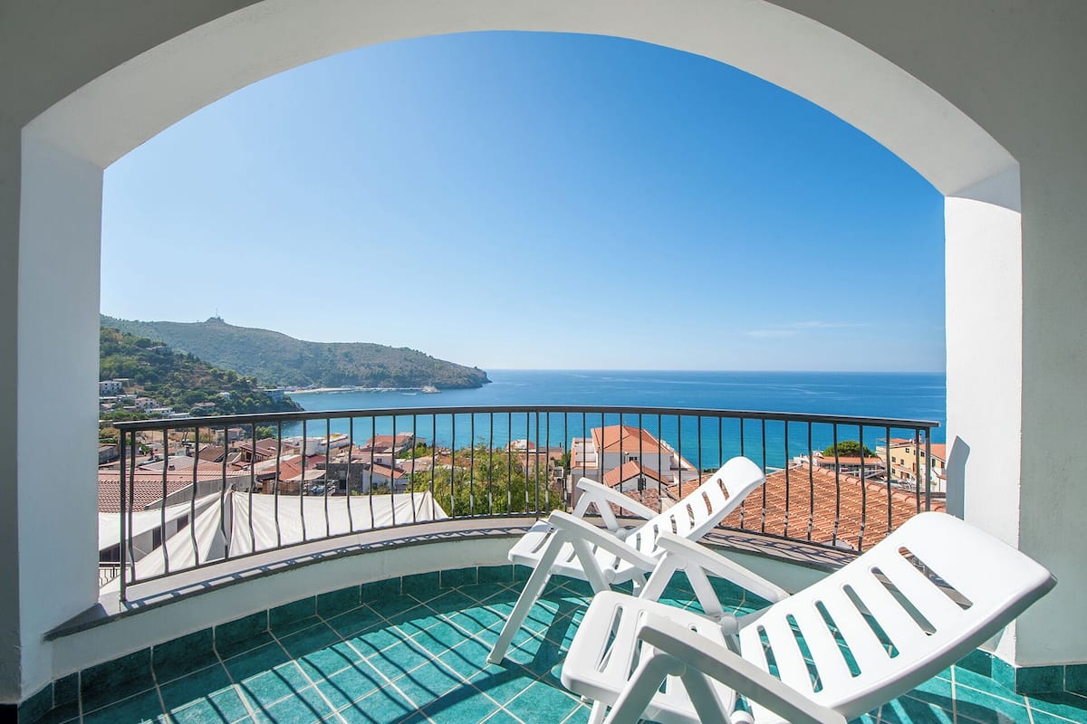 A balcony with a pool, white chairs, and a view of the sea and a hilly landscape.