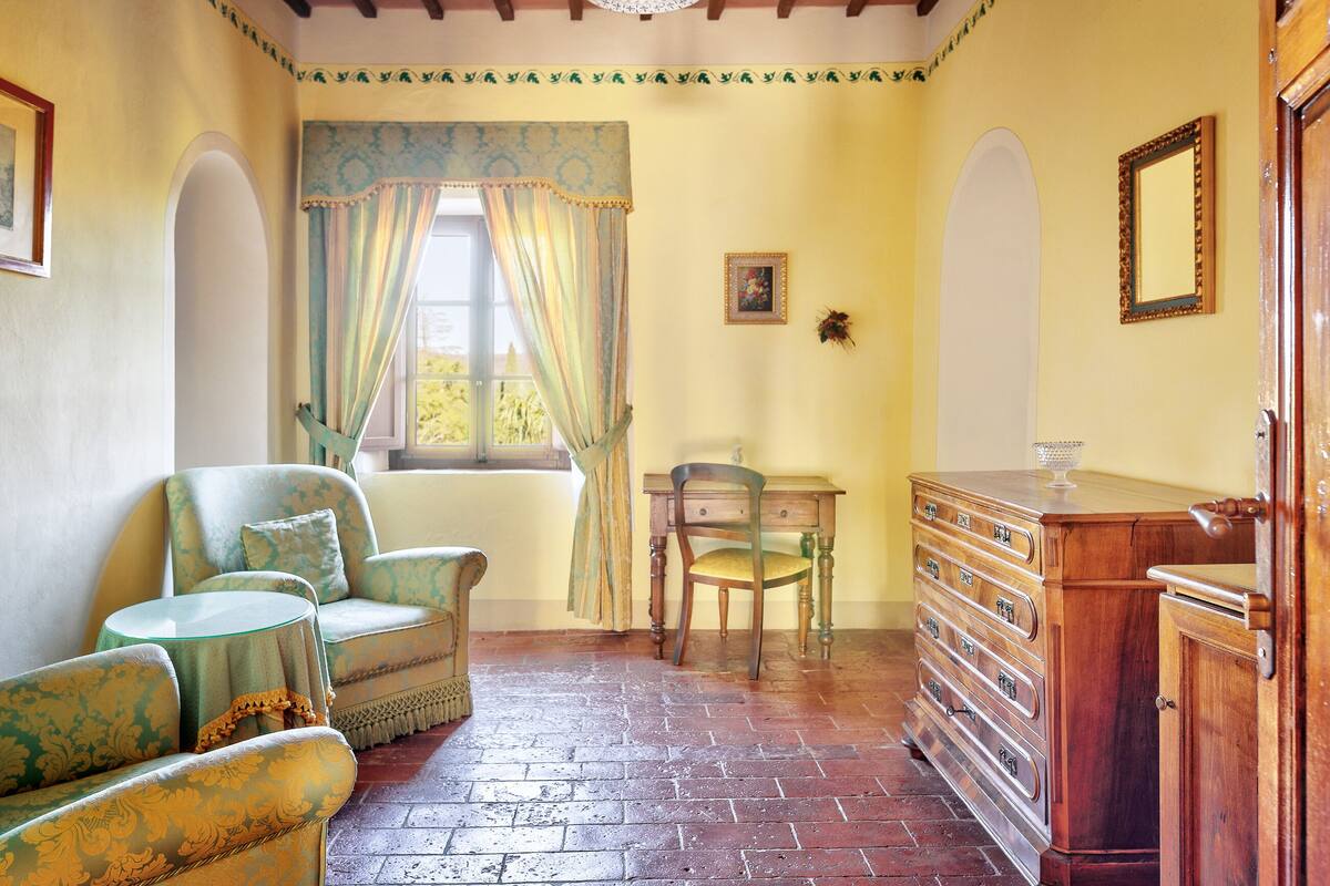 A room with a brick floor, a wooden dresser, a chair, a small table, and a window with curtains.