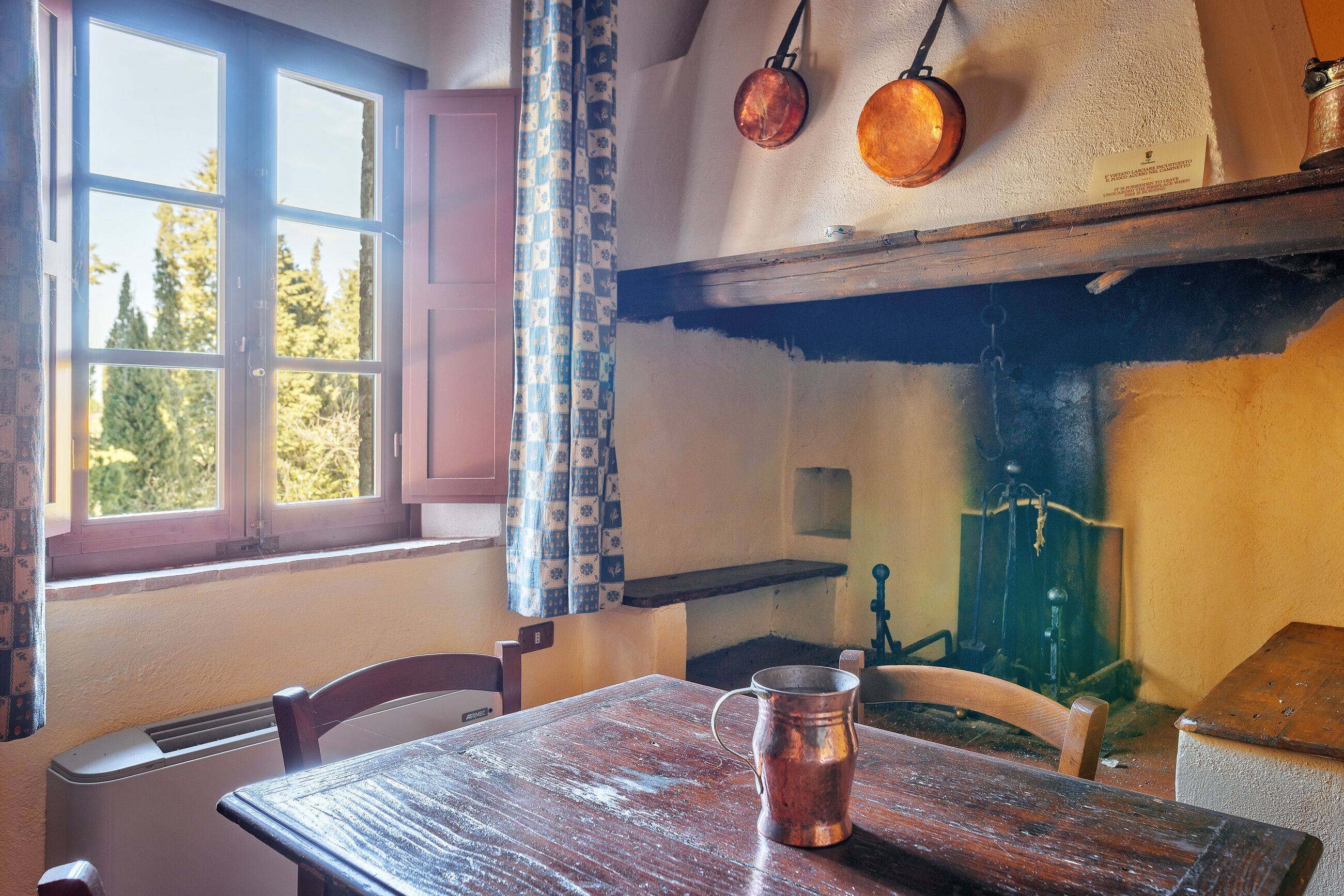Una cocina rústica con una mesa de madera, una jarra de cobre y una ventana con vista a árboles.