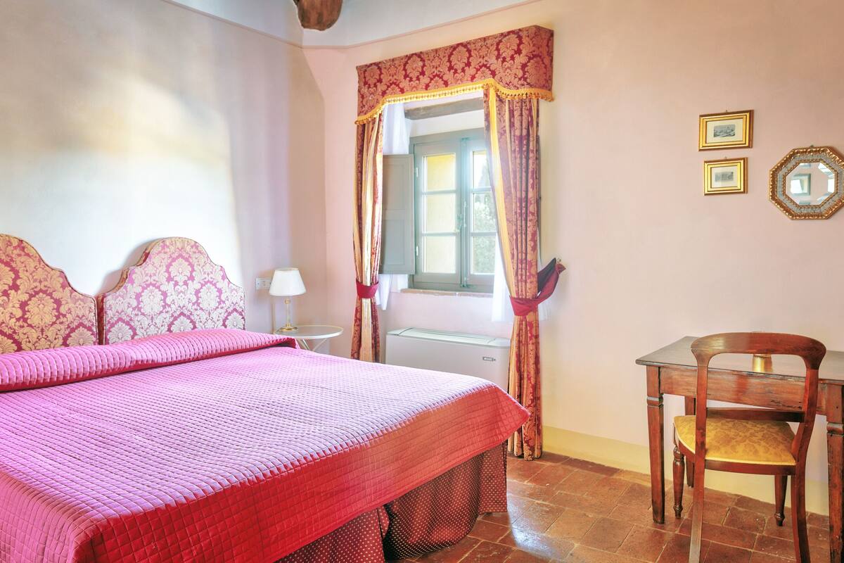 A bedroom with a bed, a chair, a table, a window with curtains, and two framed pictures on the wall.