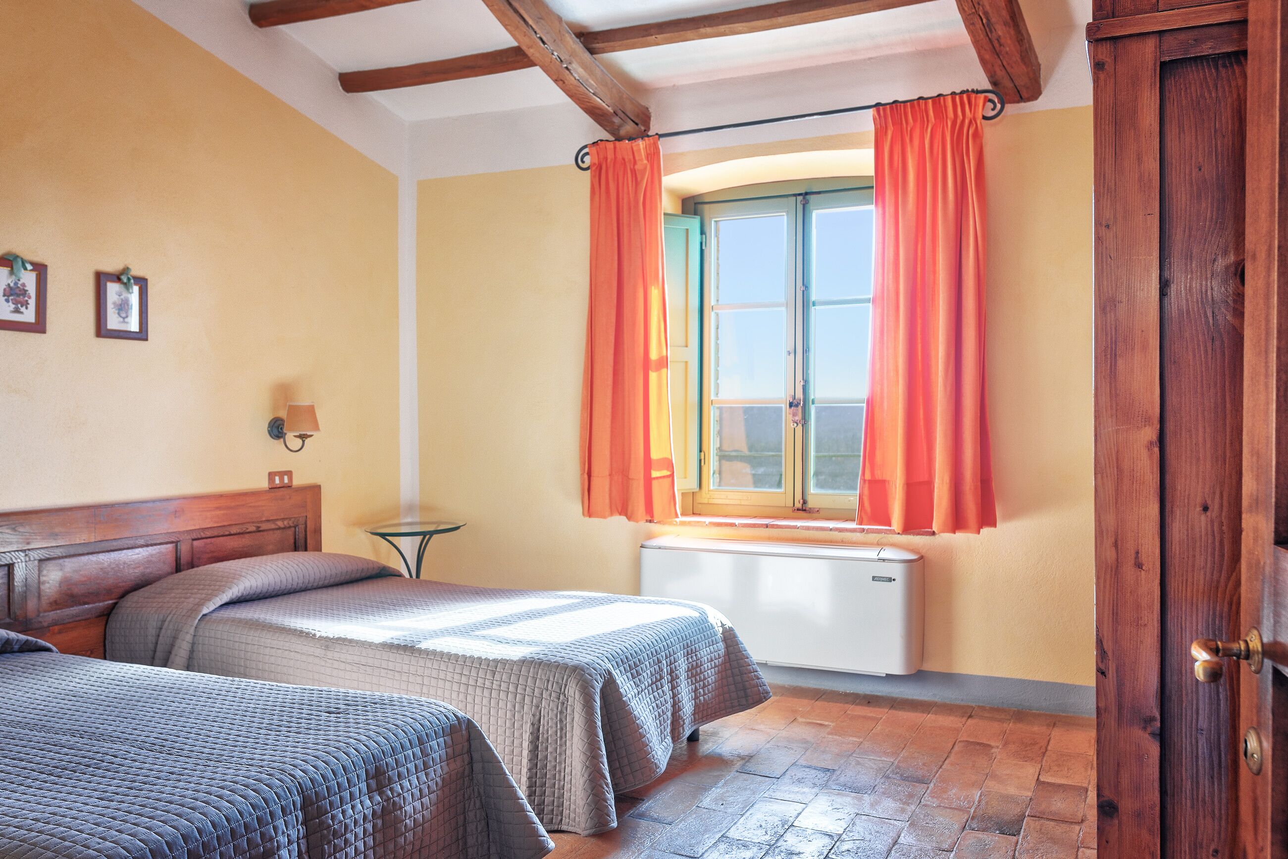 Un dormitorio con dos camas, una ventana con cortinas naranjas, un radiador y una puerta de madera.