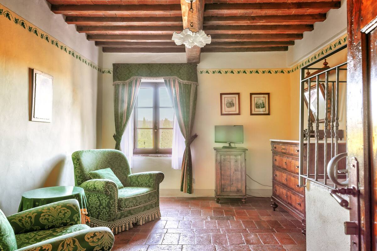 A room with a wooden ceiling, green patterned armchairs, a wooden cabinet, and a window with curtains.