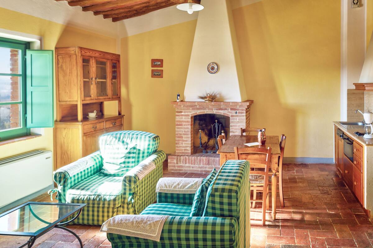 A cozy living room with a fireplace, green checkered sofas, a wooden cabinet, and a dining table.