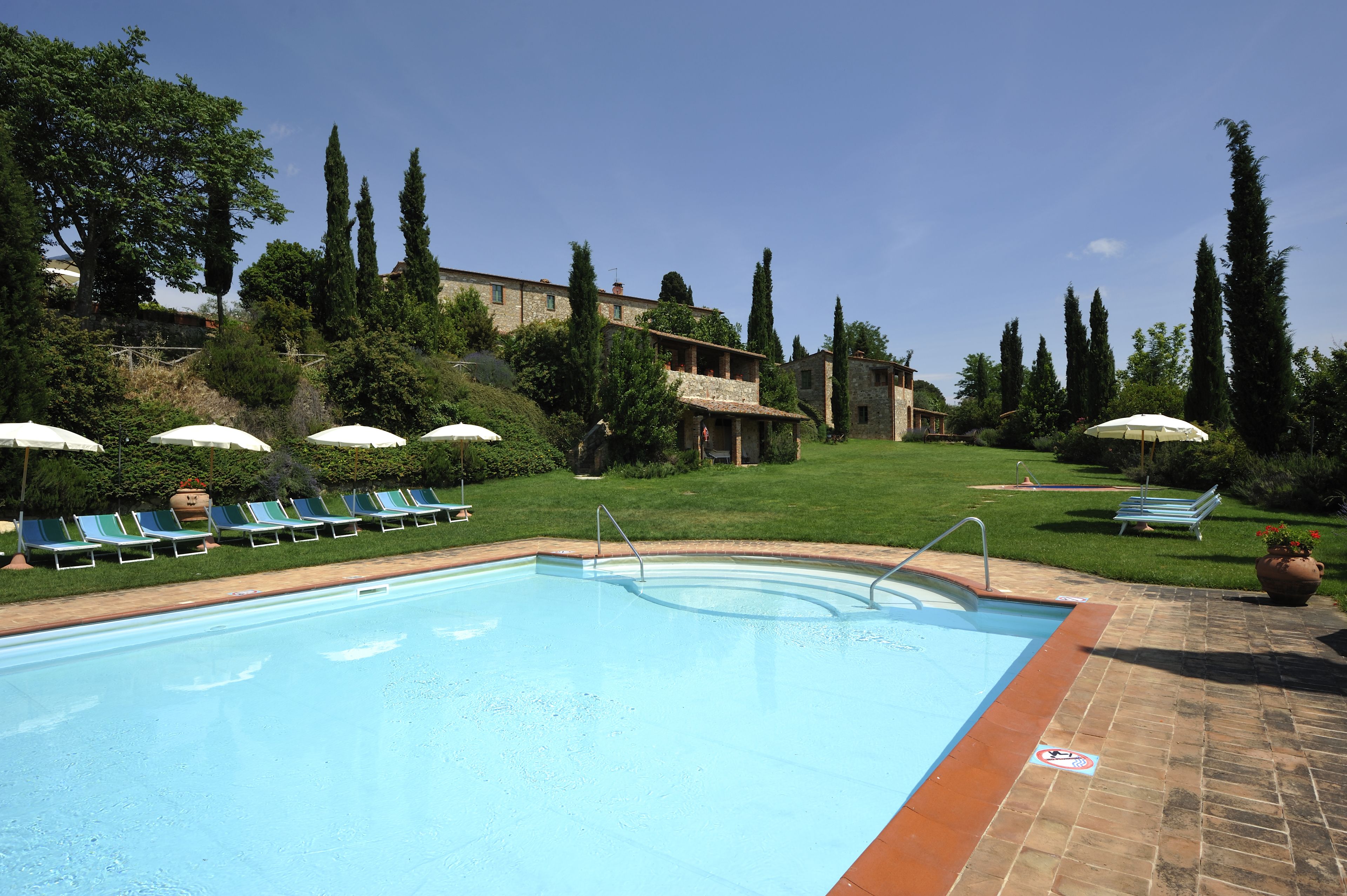 Ein Swimmingpool mit Liegestühlen und Sonnenschirmen, umgeben von Grünflächen und Gebäuden.