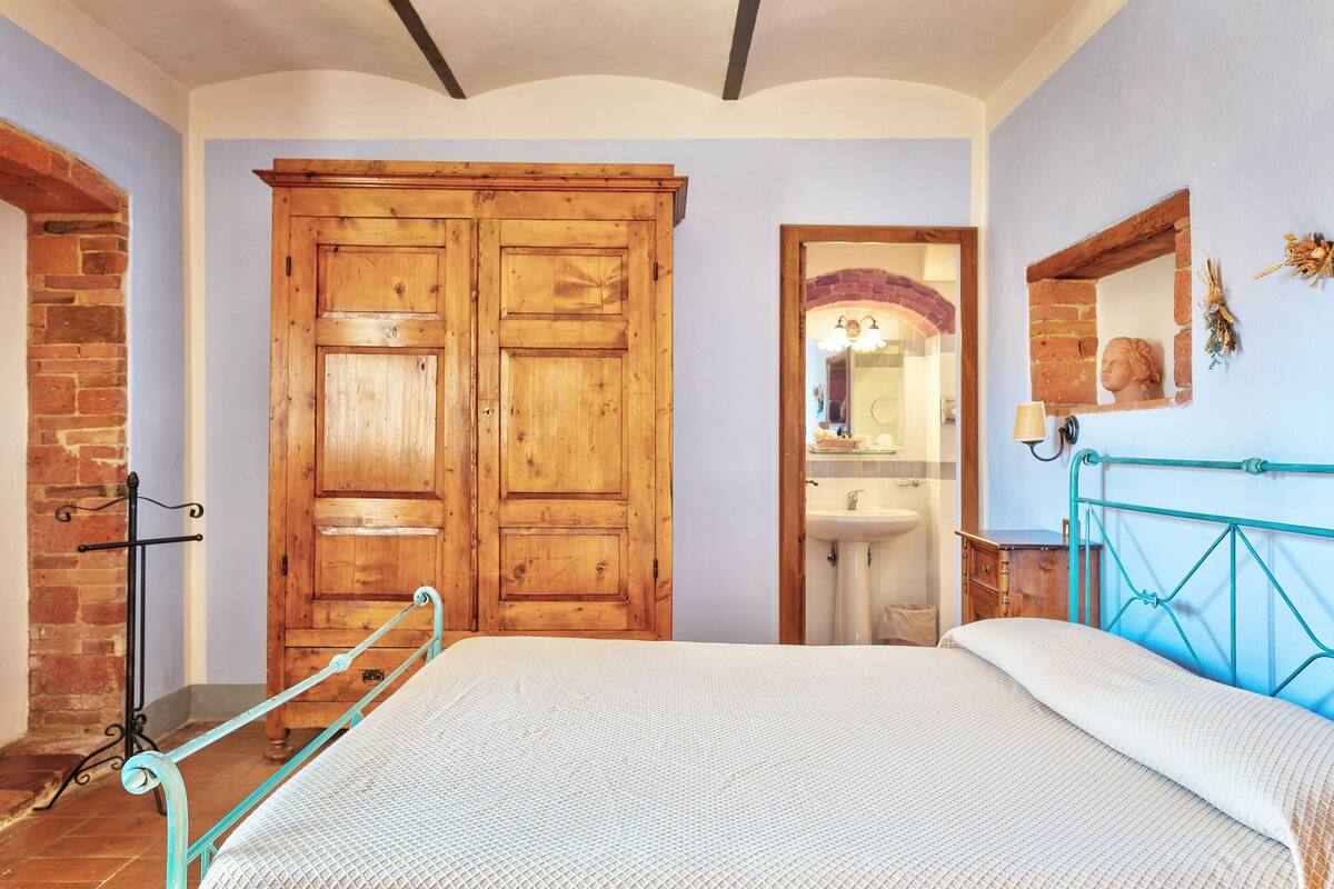 A bedroom with a wooden wardrobe, a bed with a white bedspread, a blue metal headboard, and a brick wall.