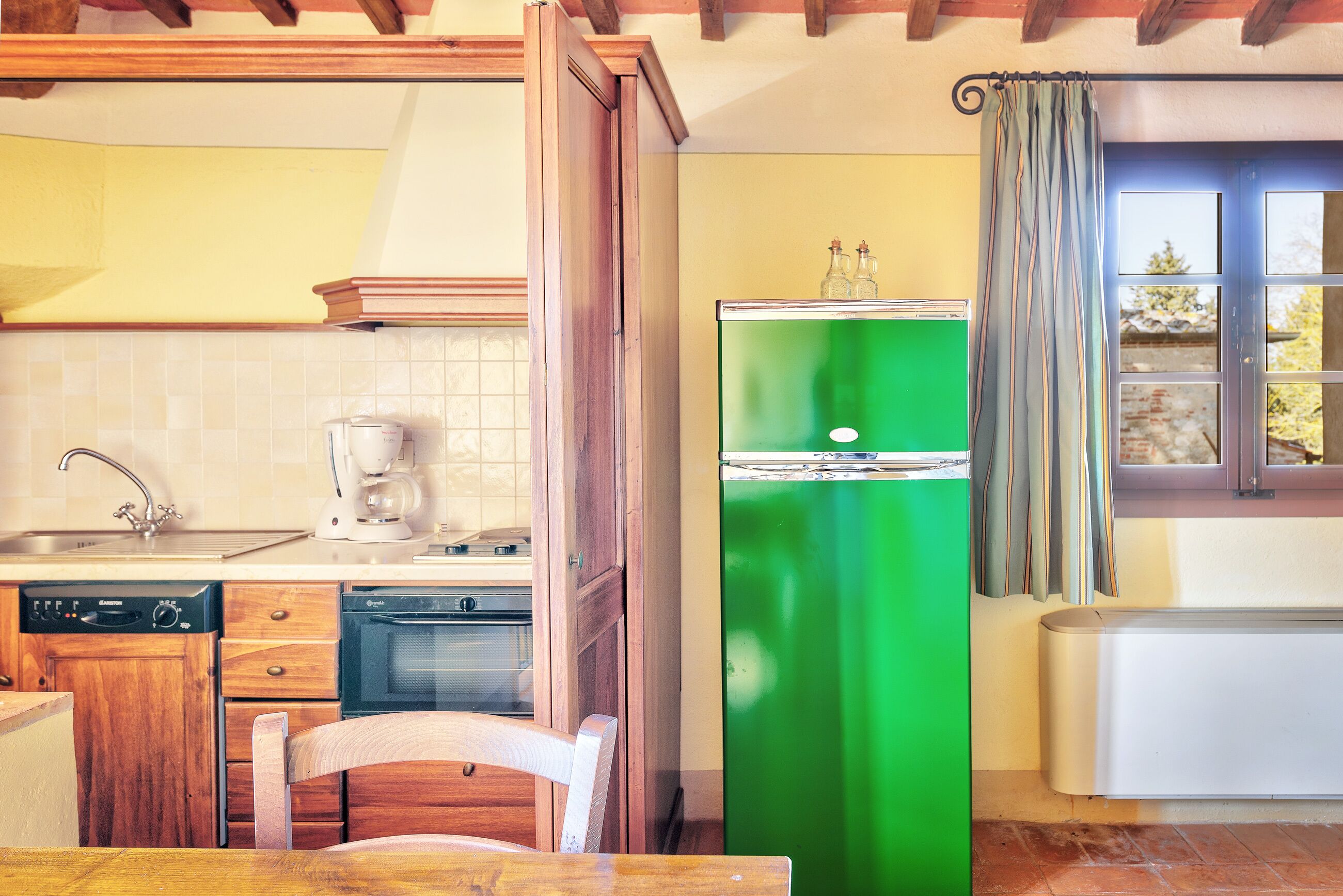 A kitchen with wooden cabinets, a green refrigerator, a coffee maker, and a window with curtains.