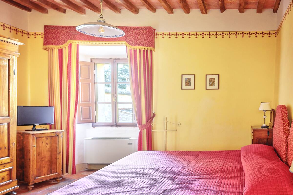 A bedroom with a large bed, a wooden dresser, a television on a cabinet, and a window with curtains.