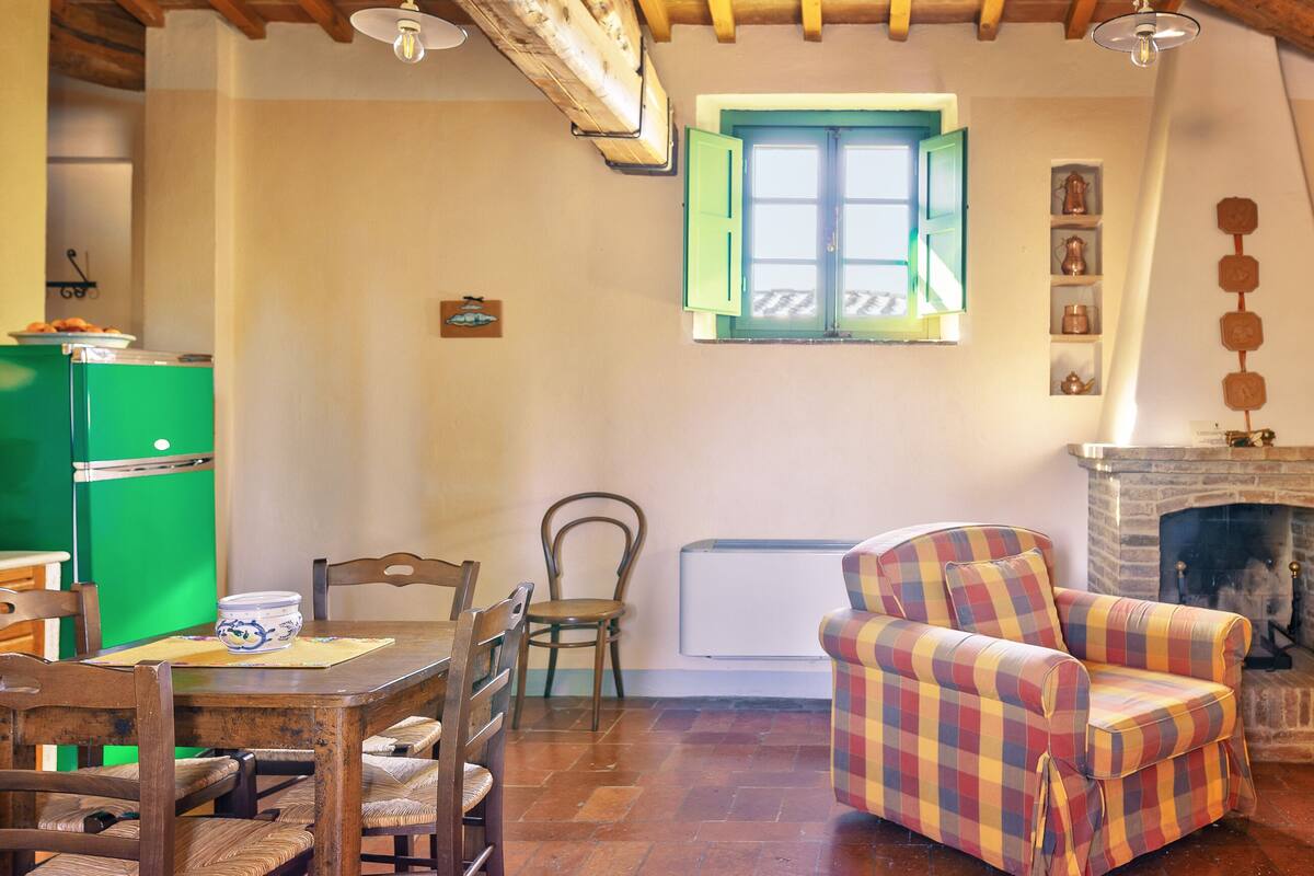A cozy living room with a dining table, chairs, a colorful armchair, a fireplace, and a window with green shutters.