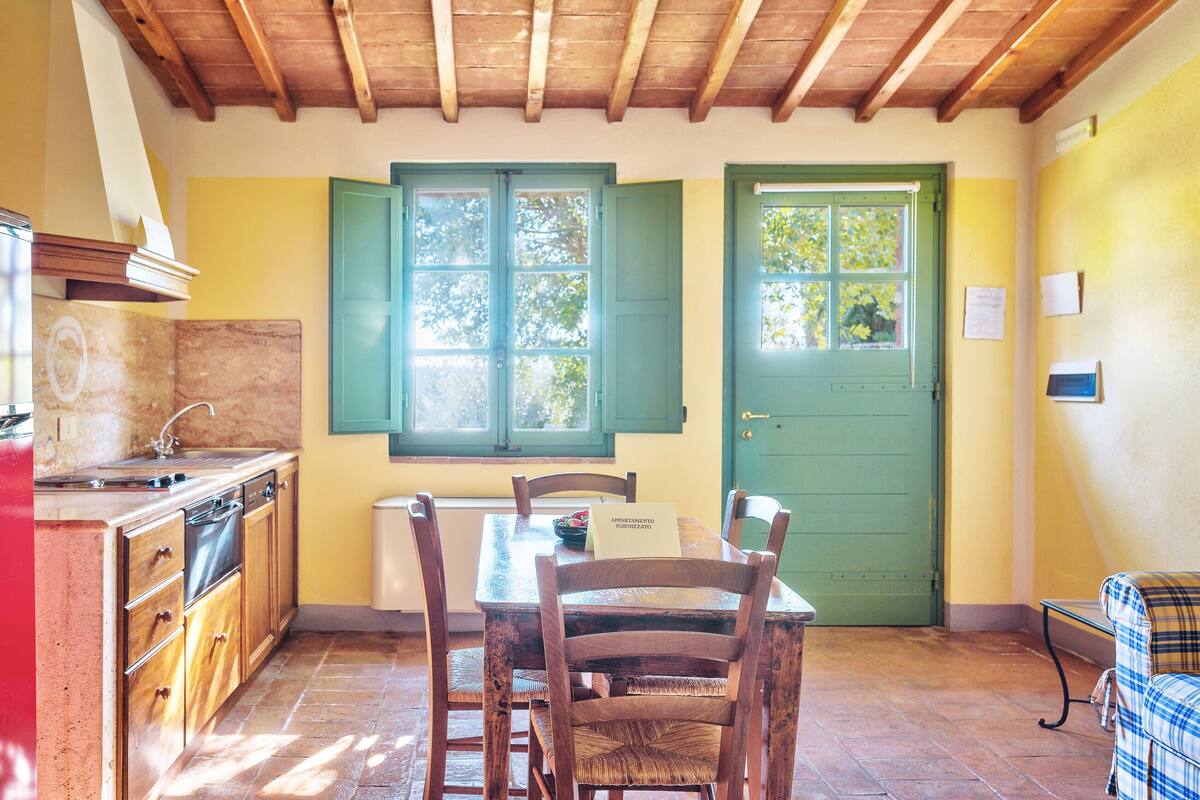 A kitchen with wooden cabinets, a dining table with chairs, a green door, and a window with shutters.