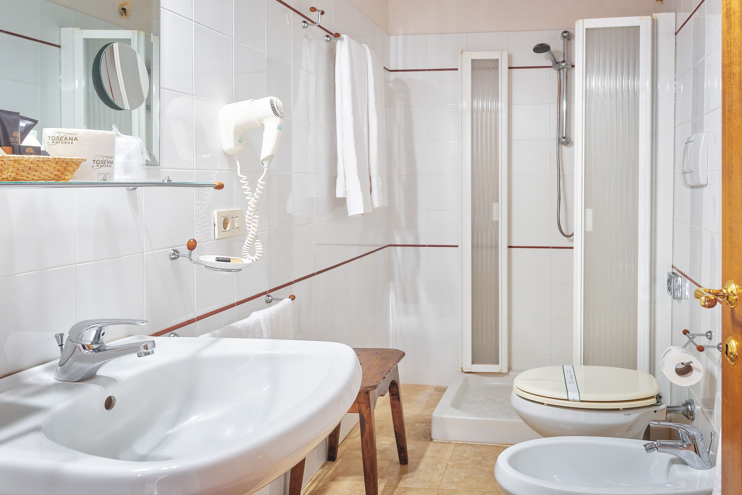 Un baño con un lavamanos blanco, un inodoro, una ducha y un taburete de madera.