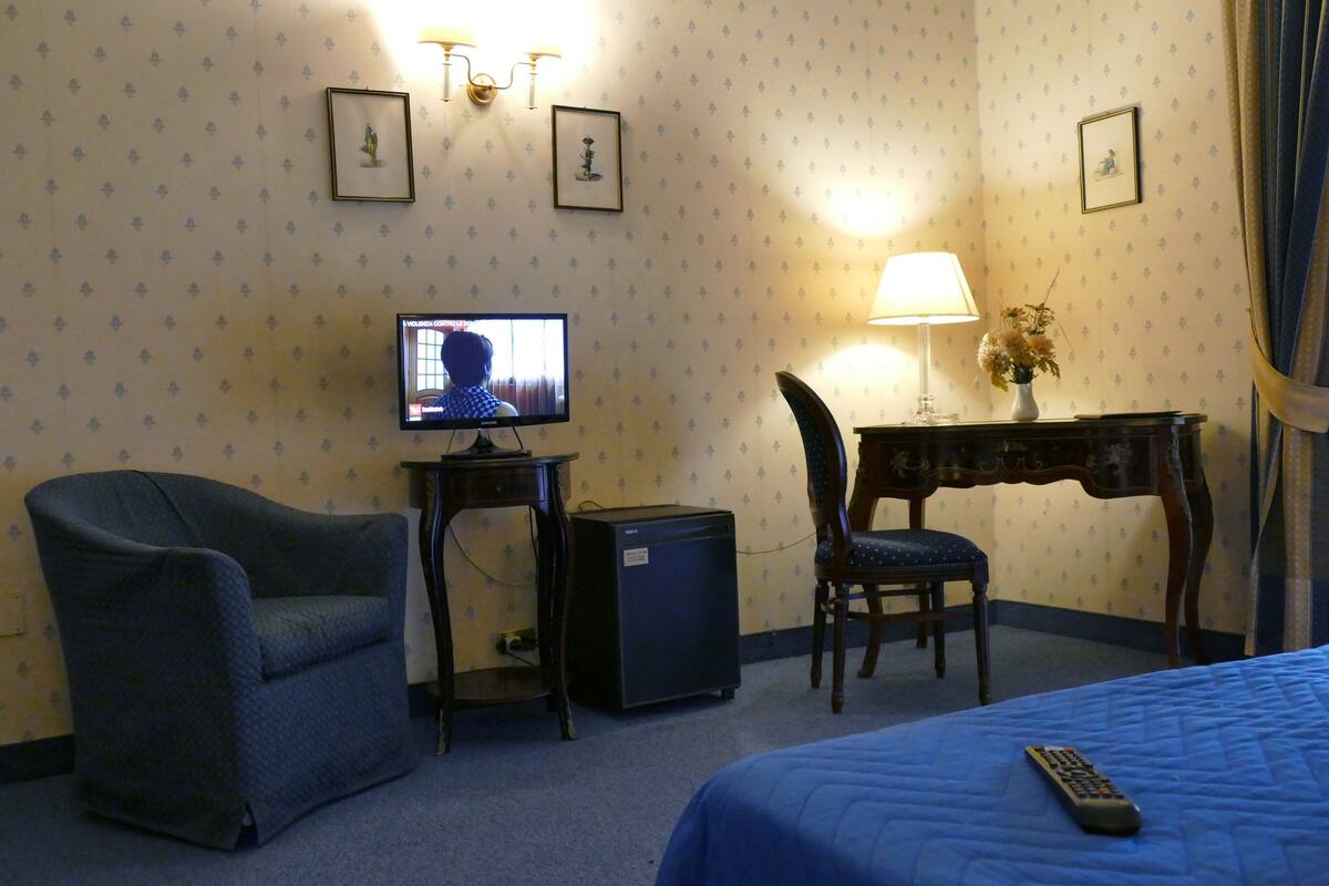 A hotel room with a bed, a chair, a desk with a lamp, a television, and a small refrigerator.