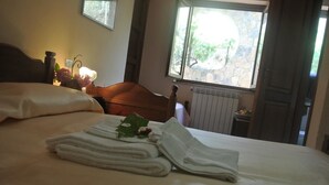 A neatly made bed with folded towels and a pair of slippers, a wooden headboard, a window with a view of greenery, and a radiator. - Il Forno Antico (San Mauro La Bruca)