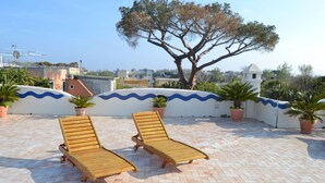Two wooden lounge chairs on a tiled patio with potted plants and a large tree in the background. - Hotel Tirreno Residence (Procida)