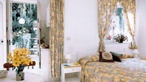 A bedroom with a bed, a bedside table, a nightstand, a chair, a window with curtains, and a hanging light fixture. - Hotel Tirreno Residence (Procida)