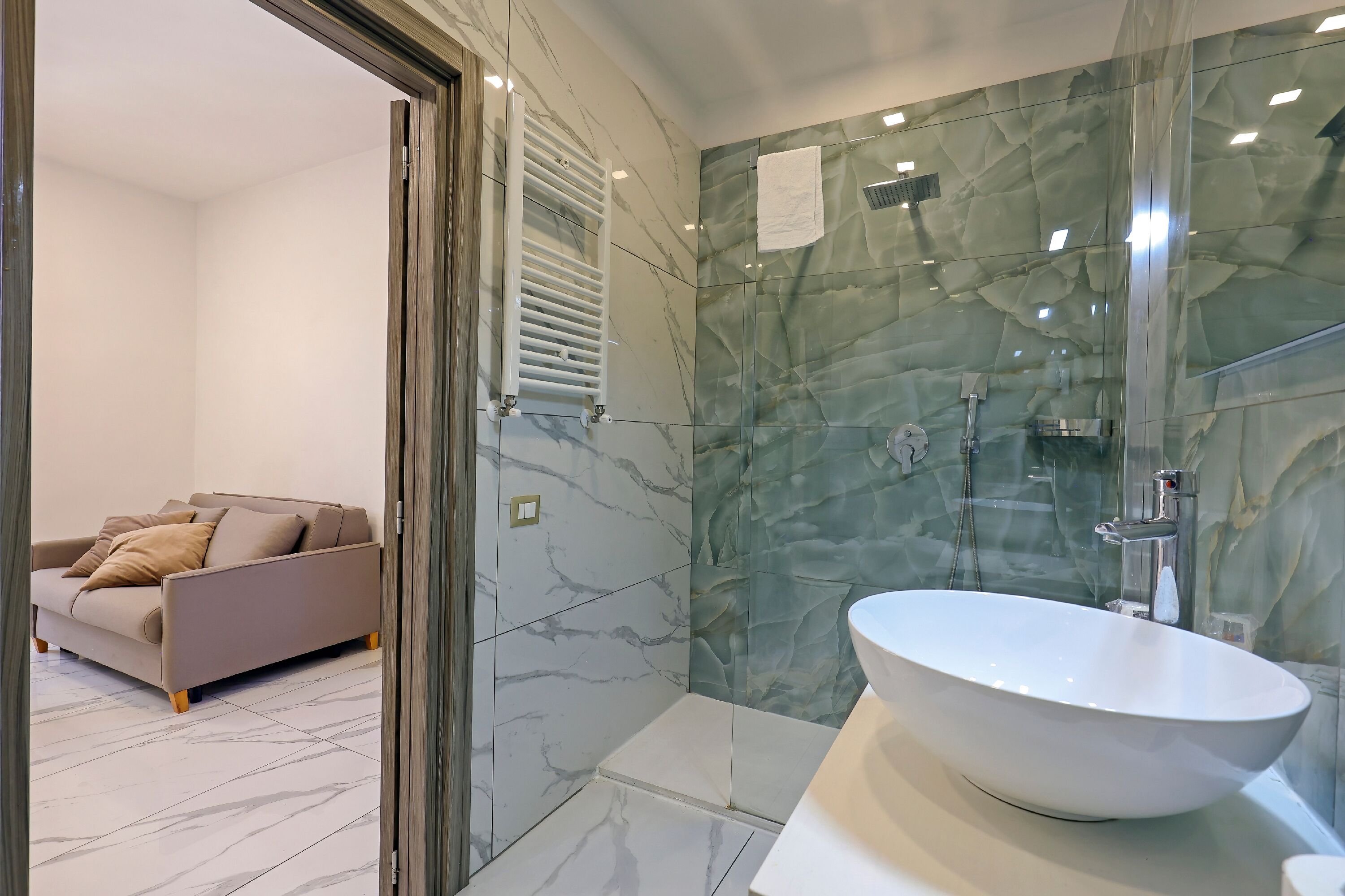 A modern bathroom with a large oval sink, a glass shower enclosure, and a beige sofa in the adjacent living area.