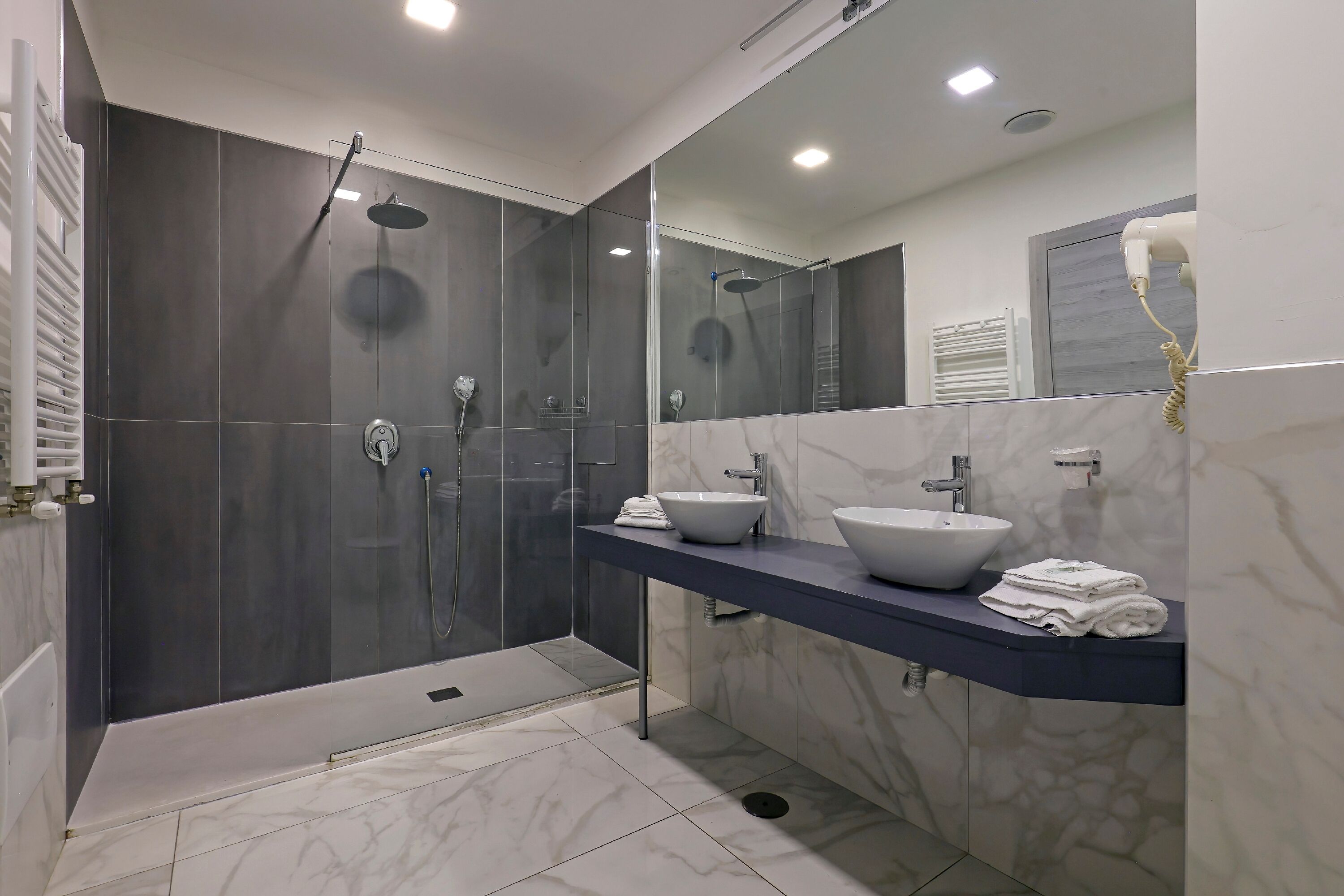 Un baño moderno con ducha a ras de suelo, tocador con dos lavamanos y secador de pelo montado en la pared.