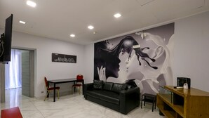 A modern living room with a black leather sofa, a wooden coffee table, and a red chair. A large wall mural depicts a woman's face. - Hotel La Pace (Naples)