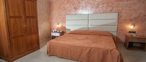 Un dormitorio con un armario grande, una cama con cabecera, dos mesitas de noche con lámparas y una colcha estampada.