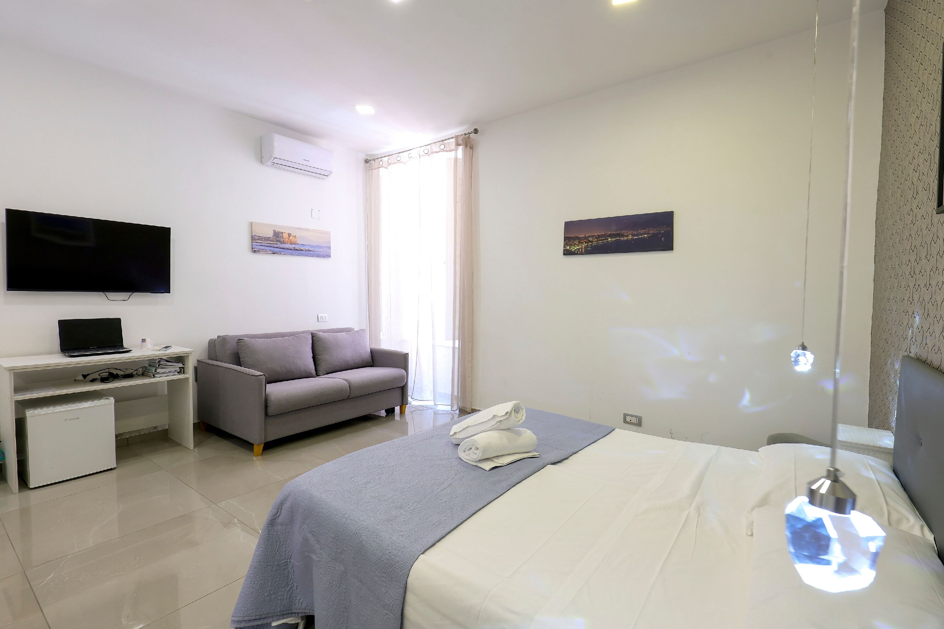 A hotel room with a bed, a desk with a laptop, a sofa, and a TV.