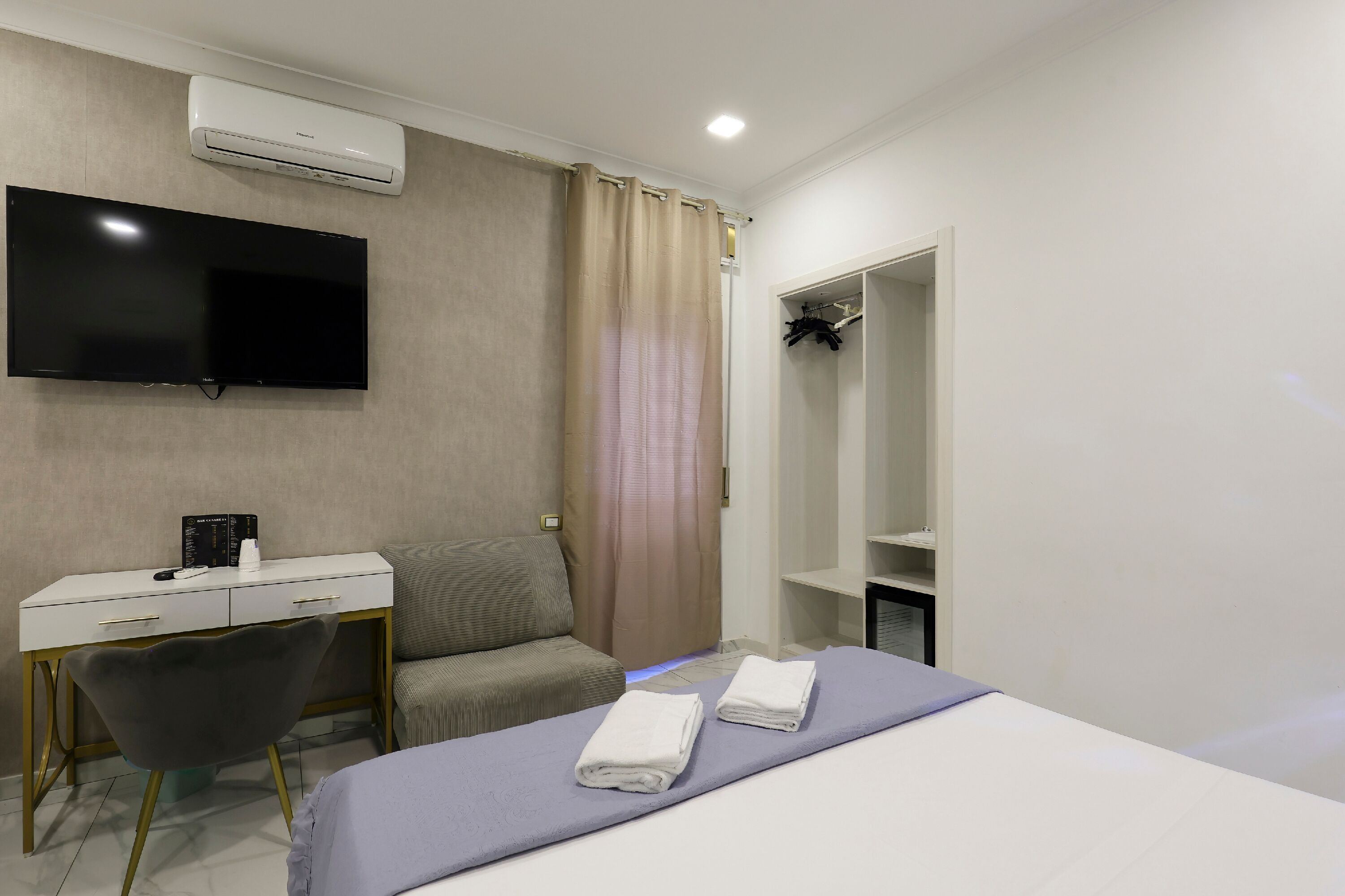 Una habitación de hotel con cama, escritorio con silla, televisor montado en la pared, armario y cortina de ducha.
