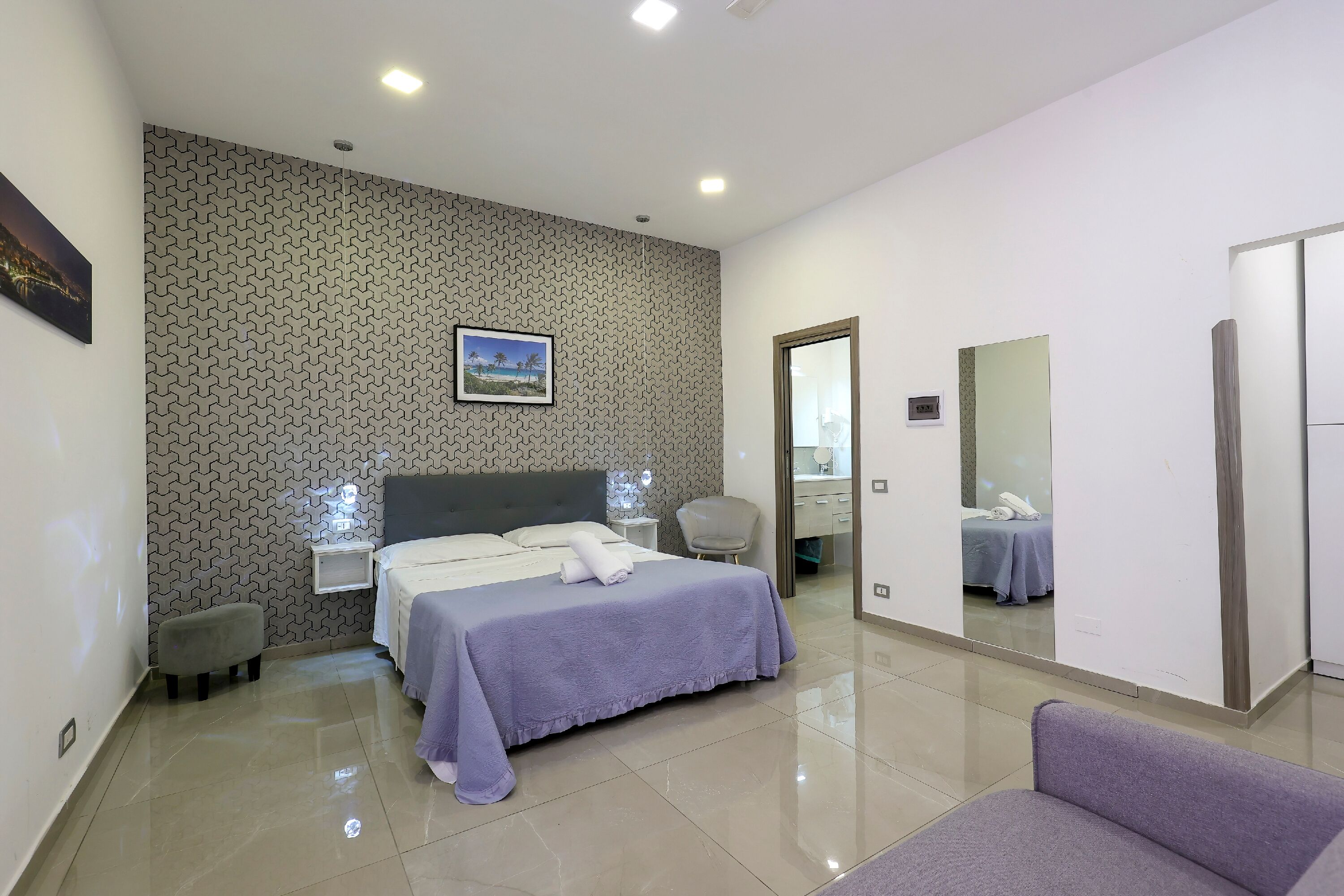 A modern hotel room with a neatly made bed, a small stool, a chair, and a large mirror.