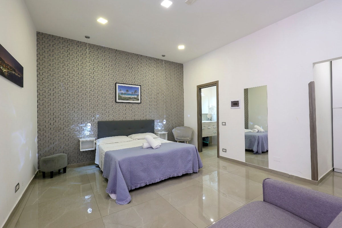 Una camera d'hotel moderna con un letto rifatto, uno sgabello, una sedia e uno specchio grande.