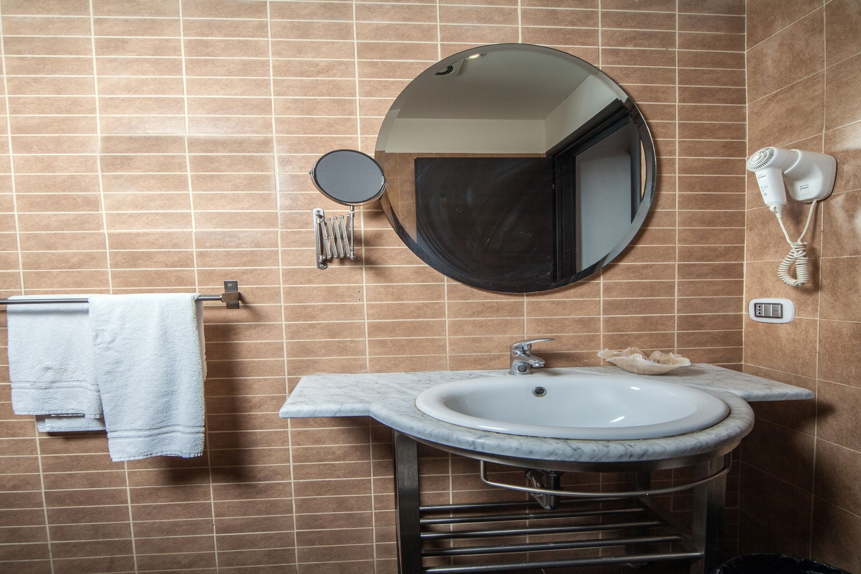 A bathroom with a round mirror, a sink, a towel rack, and a hairdryer mounted on the wall.