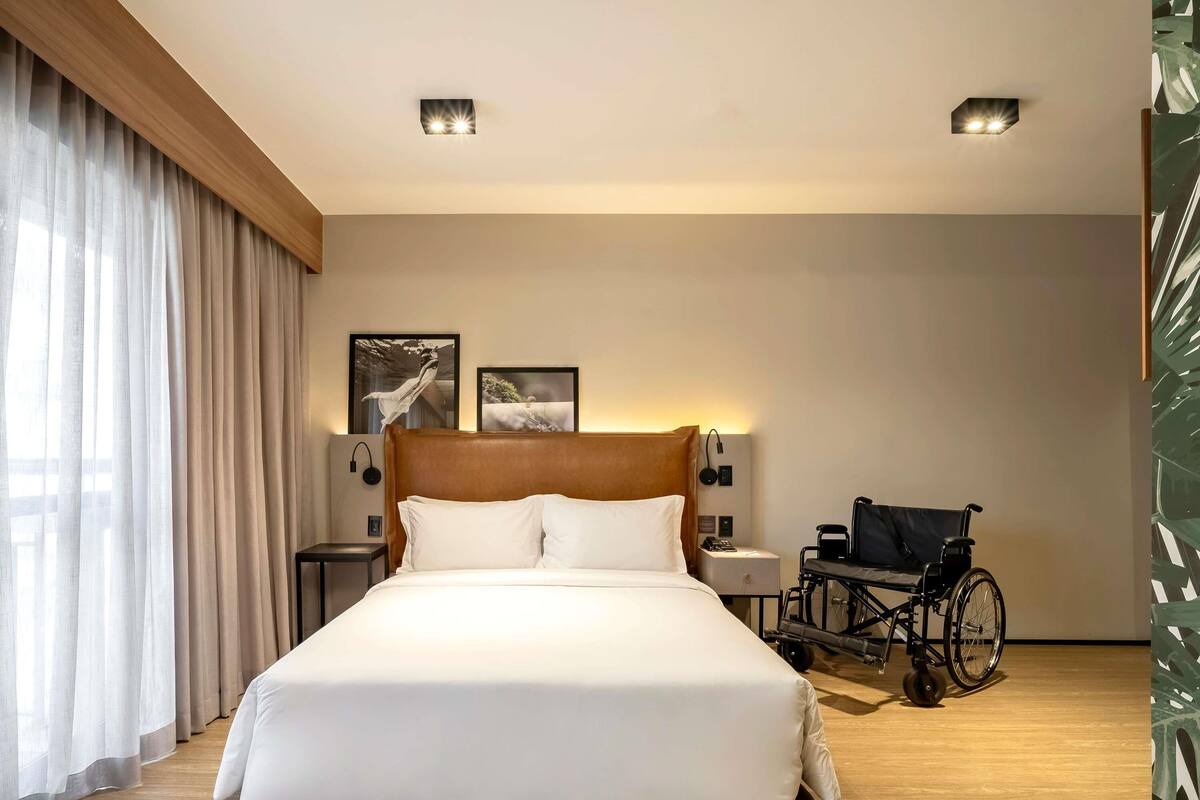 Et hotelværelse med en stor seng, en kørestol, et natbord og to vægmonterede lamper.