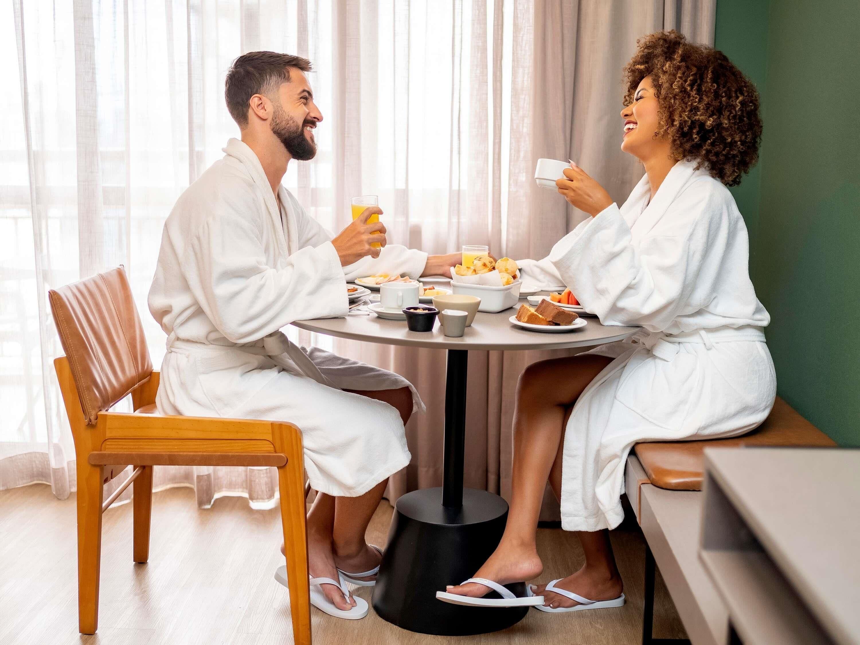 A couple in white robes at a round dining table with breakfast items, a wooden chair, and a black floor lamp.