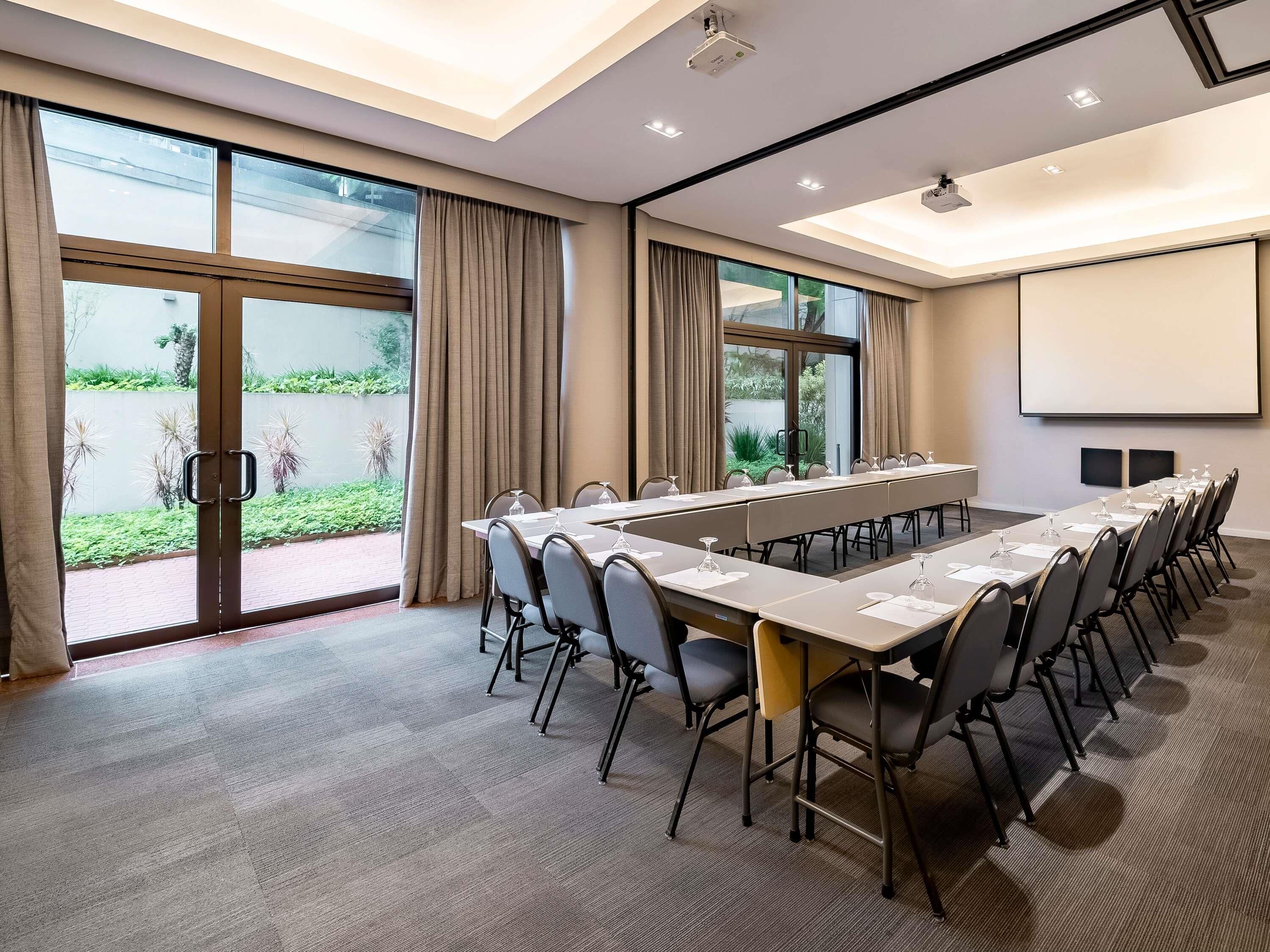 A conference room with a long table, chairs, a projector screen, and large windows with curtains.