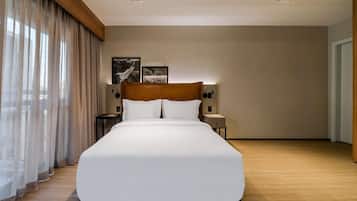 A large bed with a wooden headboard in a hotel room. There are two bedside tables with lamps, a large window with curtains, and two wall-mounted light fixtures.