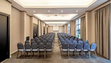 Uma sala de conferências com fileiras de cadeiras, uma tela de projetor e grandes janelas.