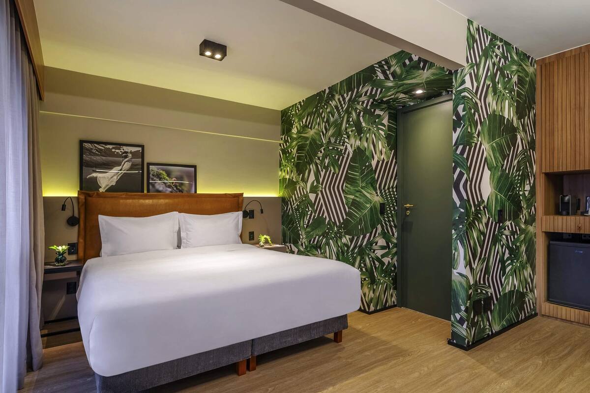 Et moderne hotelværelse med en stor seng, et træhovedgærde, et sengebord med en lampe og en væg med et tropisk bladmotiv.