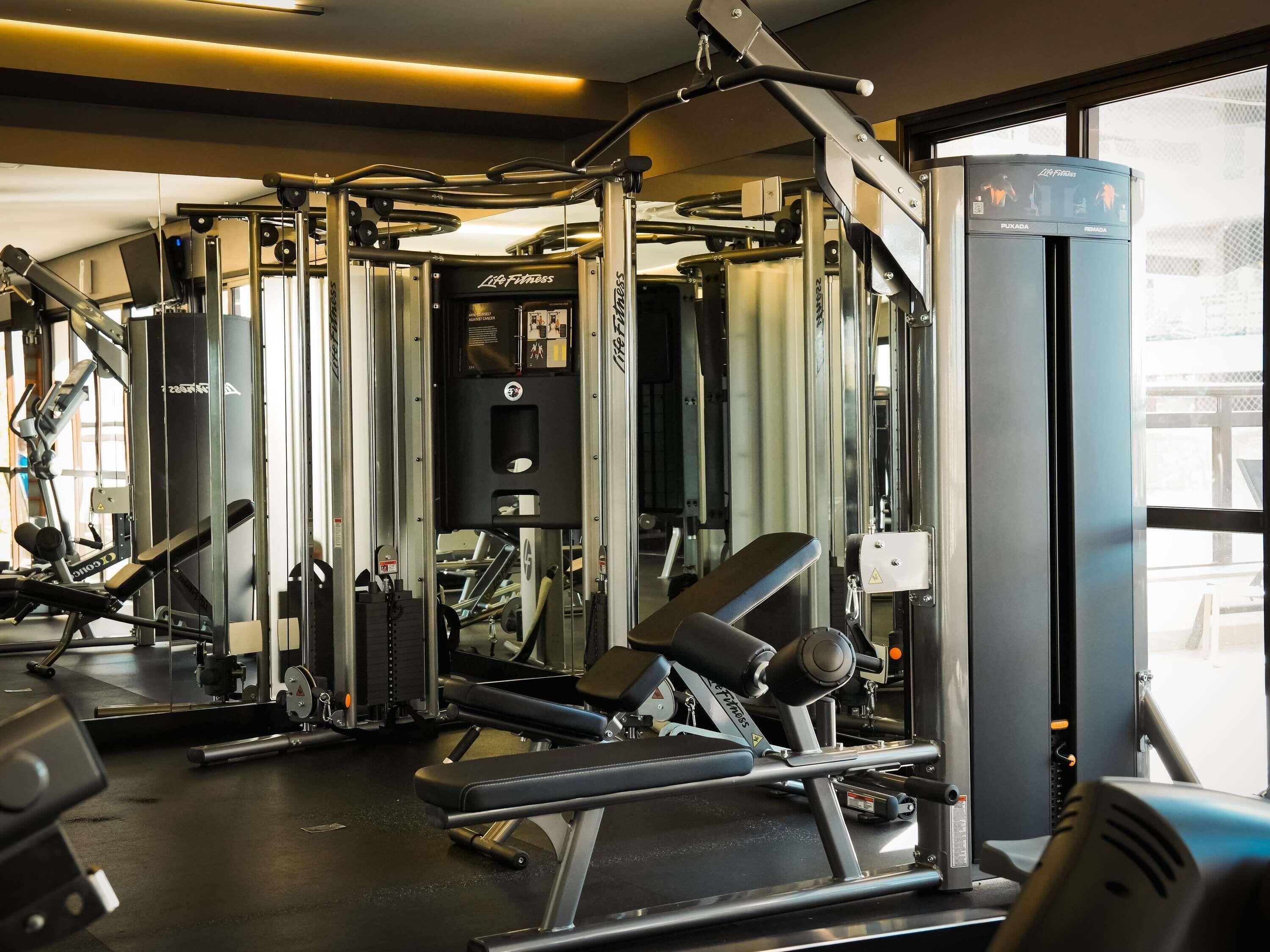 A gym with various exercise machines, including a Smith machine and a cable machine, with a window providing natural light.