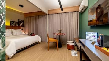 A hotel room with a bed, a desk, a chair, a table with a drink, and a painting on the wall.