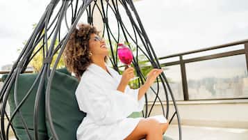 A person sitting on a green cushioned chair in a wicker chair suspended from the ceiling, holding a pink drink with a garnish.