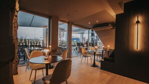 A modern dining area with round tables and chairs, large windows offering a city view, and a cozy seating area with a sofa and lamp. - Bisonte Libertad (Buenos Aires)