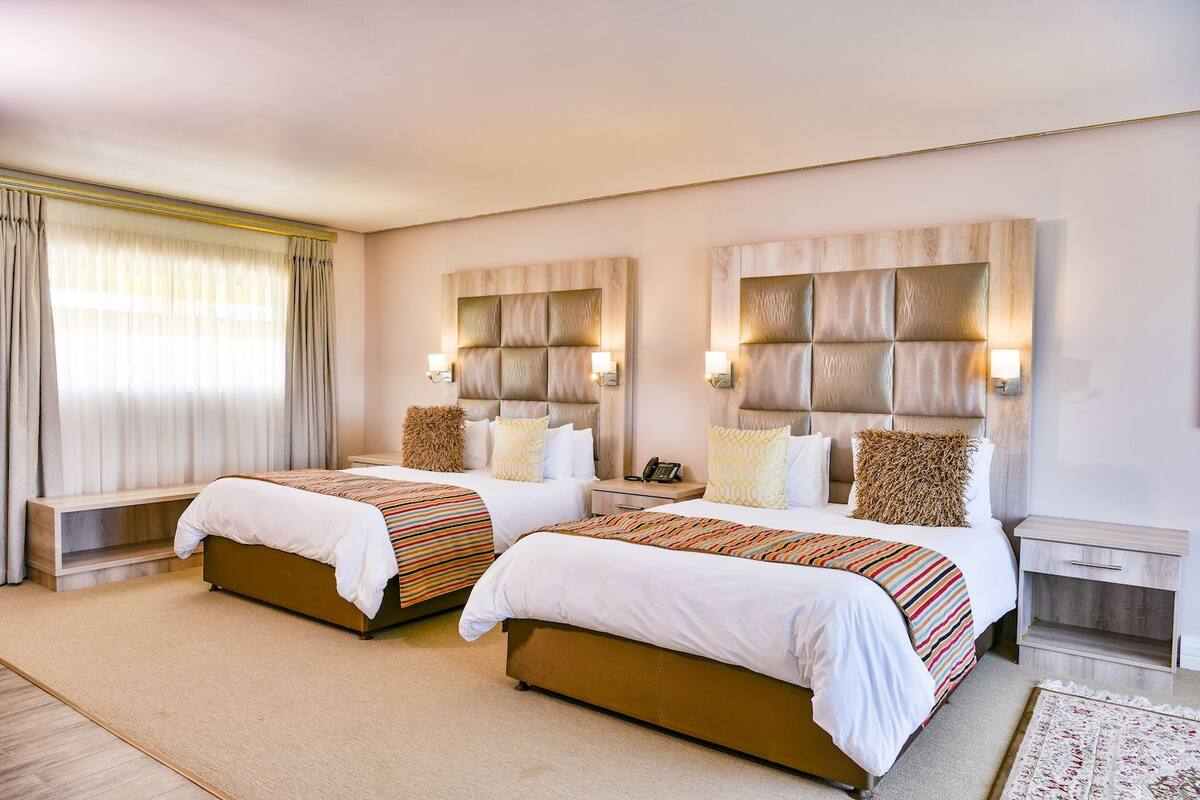 Une chambre d’hôtel avec deux lits, une grande fenêtre avec des rideaux, une table de chevet avec une lampe et un petit tapis.