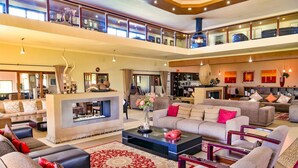 A spacious, well-lit living area with a fireplace, multiple seating options, and a balcony with luggage. - Kwantu Private Game Reserve (Sidbury)
