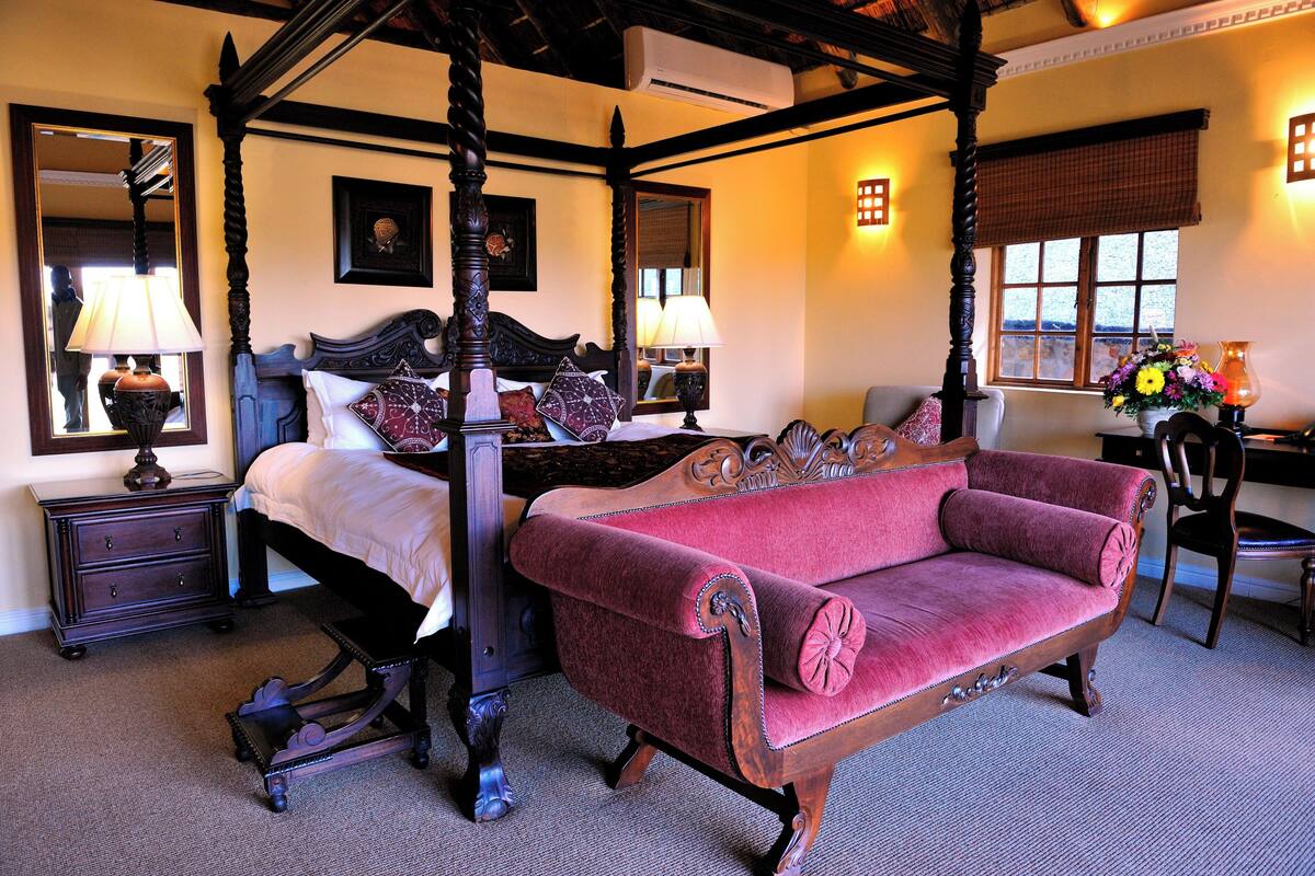 Un lit à baldaquin avec un canapé rouge recouvert de tissu, une table de chevet, un miroir et une fenêtre donnant sur un paysage.