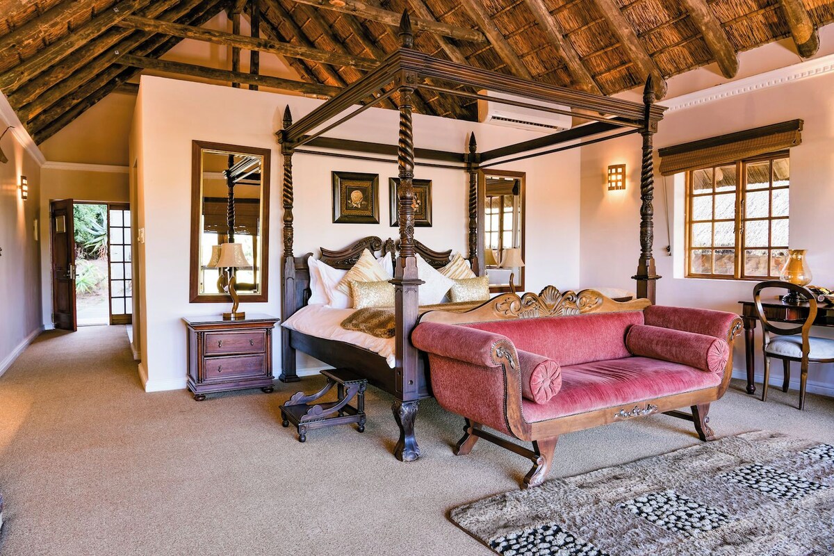 A four-poster bed, a chaise lounge, a nightstand, a desk, and a chair in a room with a wooden ceiling and a large window.
