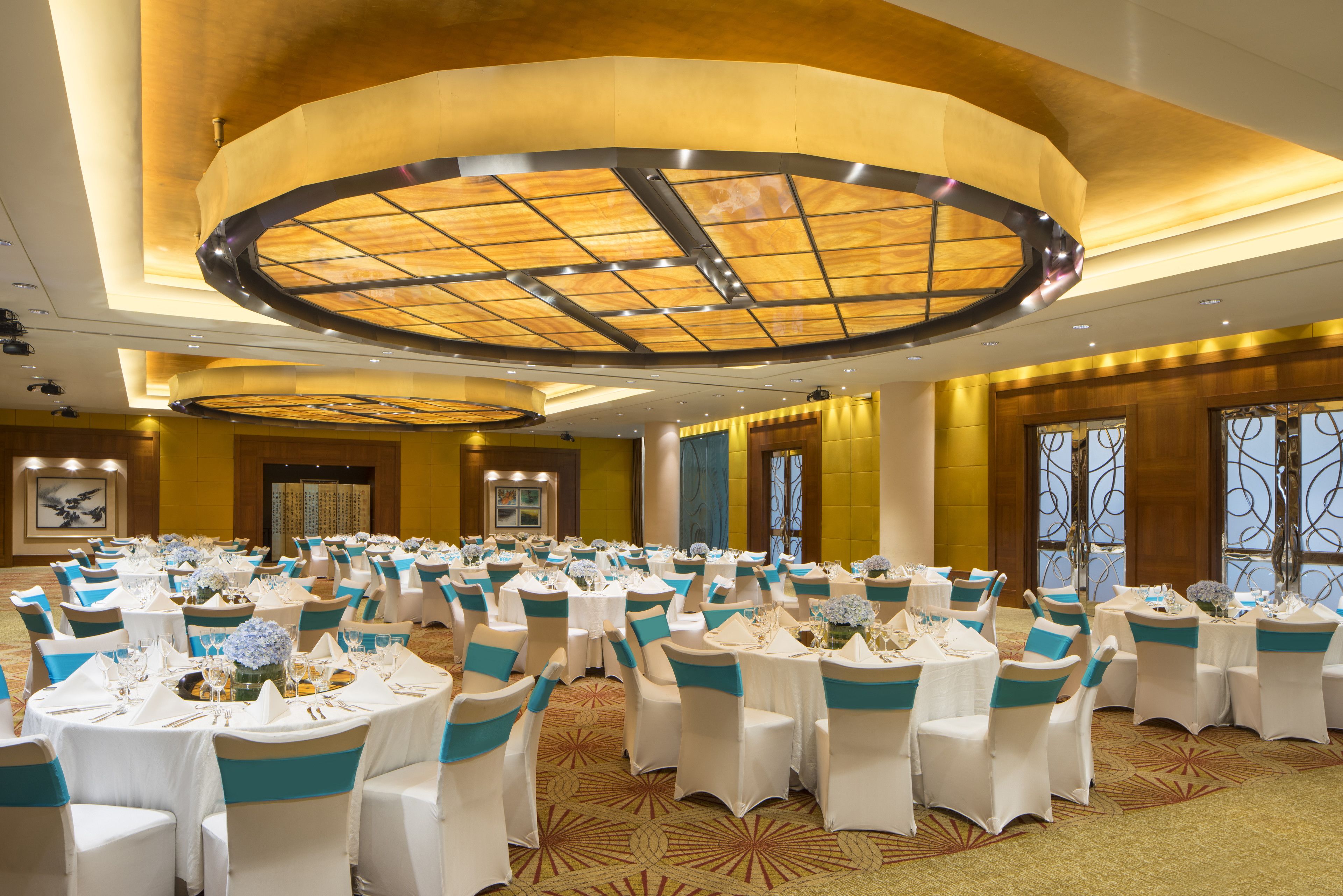 Une salle de banquet avec des tables rondes, des chaises blanches ornées de rubans bleus et un plafond au design moderne.