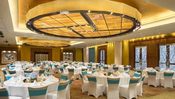 Une salle de banquet avec des tables rondes, des chaises blanches ornées de rubans bleus et un plafond au design moderne.